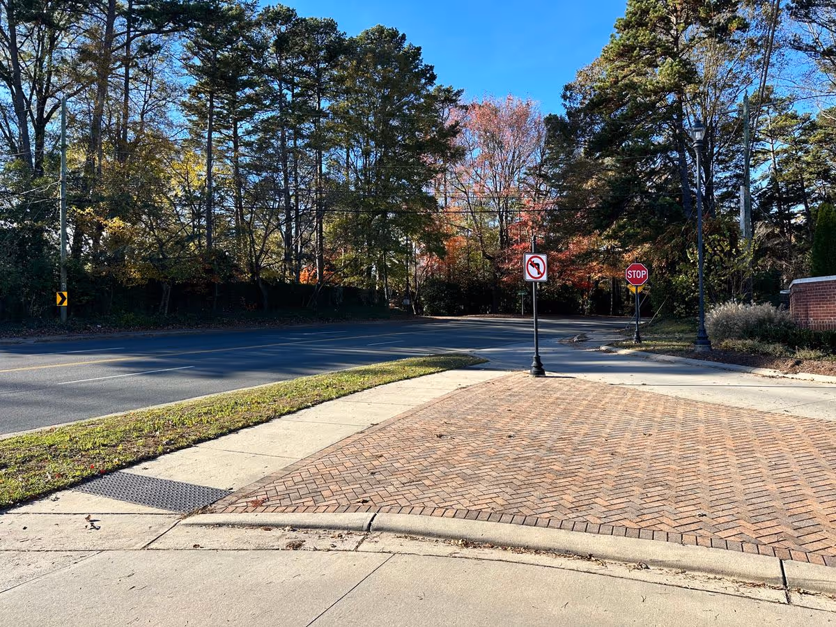 Brick-paved driveway and sidewalk at a tree-lined street intersection with a stop sign and no-left-turn sign.