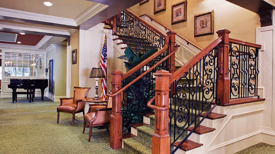 Interior view of a senior living facility featuring a carpeted staircase with ornate wooden handrails and black wrought iron balusters. Two upholstered chairs and a small round table with a lamp are placed near the staircase. An American flag stands in the corner, and a black grand piano is visible in the background near a large window. The walls are decorated with framed artwork.