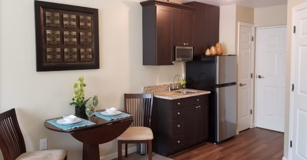 Small kitchen area with dark wood cabinets, a granite countertop with a sink, a microwave above the counter, and a stainless steel refrigerator. Next to the kitchen is a small dining table set for two with plates, napkins, and wine glasses. A decorative framed artwork hangs on the wall above the table. The floor is wooden, and there are white doors visible in the background.