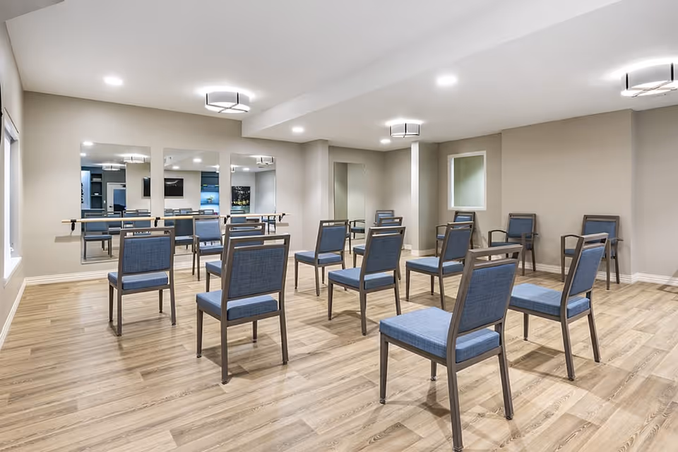 A spacious room with wooden flooring and beige walls, furnished with multiple blue cushioned chairs arranged in rows facing a wall with large mirrors. The room is well-lit with ceiling lights and has a ballet barre attached to the mirrored wall.