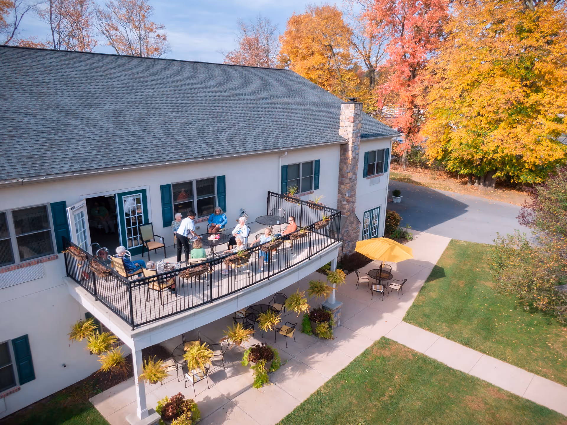 Second-floor outdoor terrace at a senior living facility with people seated around tables, a lower patio with an umbrella, and colorful autumn trees behind the building.