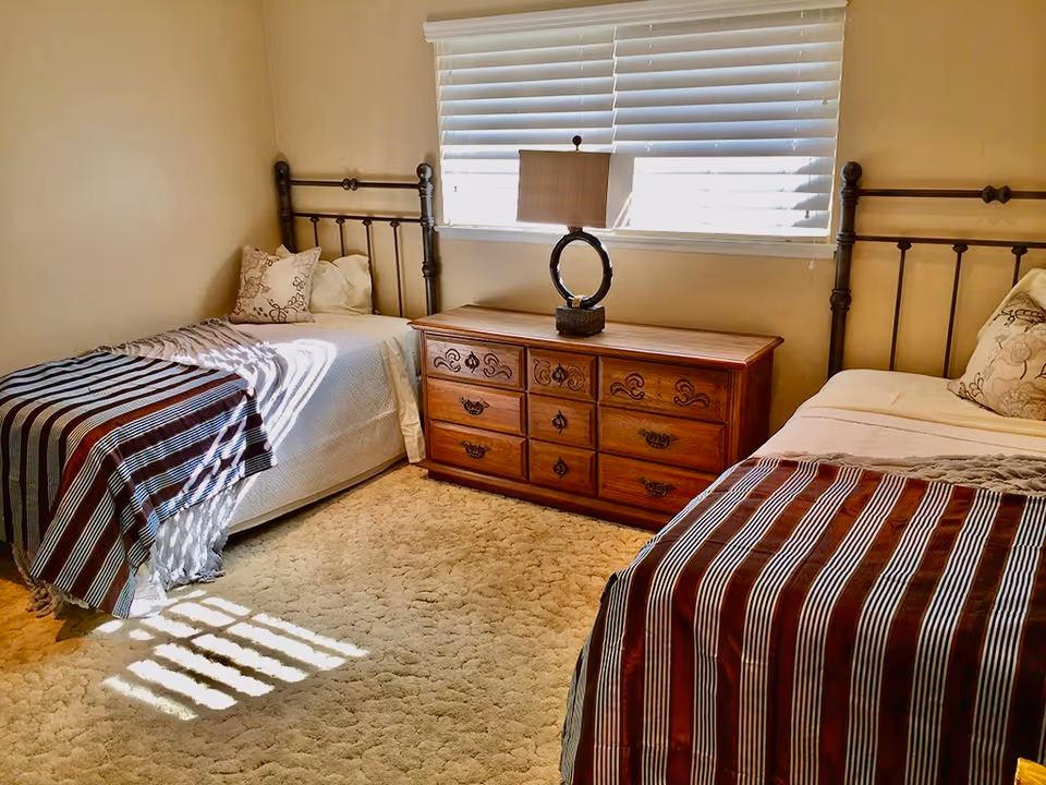 Bright bedroom with two twin beds, a wooden dresser and lamp between them beneath a window with blinds.