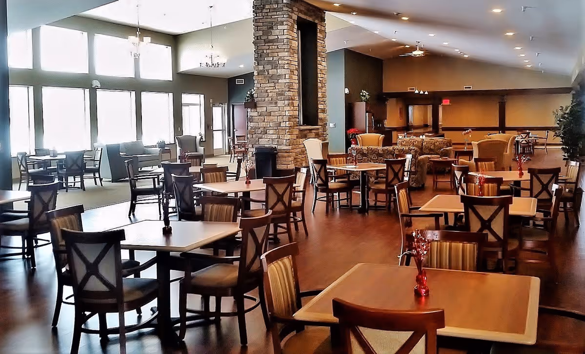 Spacious dining room with multiple wooden tables and chairs arranged neatly. The room features large windows letting in natural light, a stone pillar in the center, and comfortable seating areas with sofas and armchairs in the background. The floor is wooden, and small red vases with decorative elements are placed on the tables.