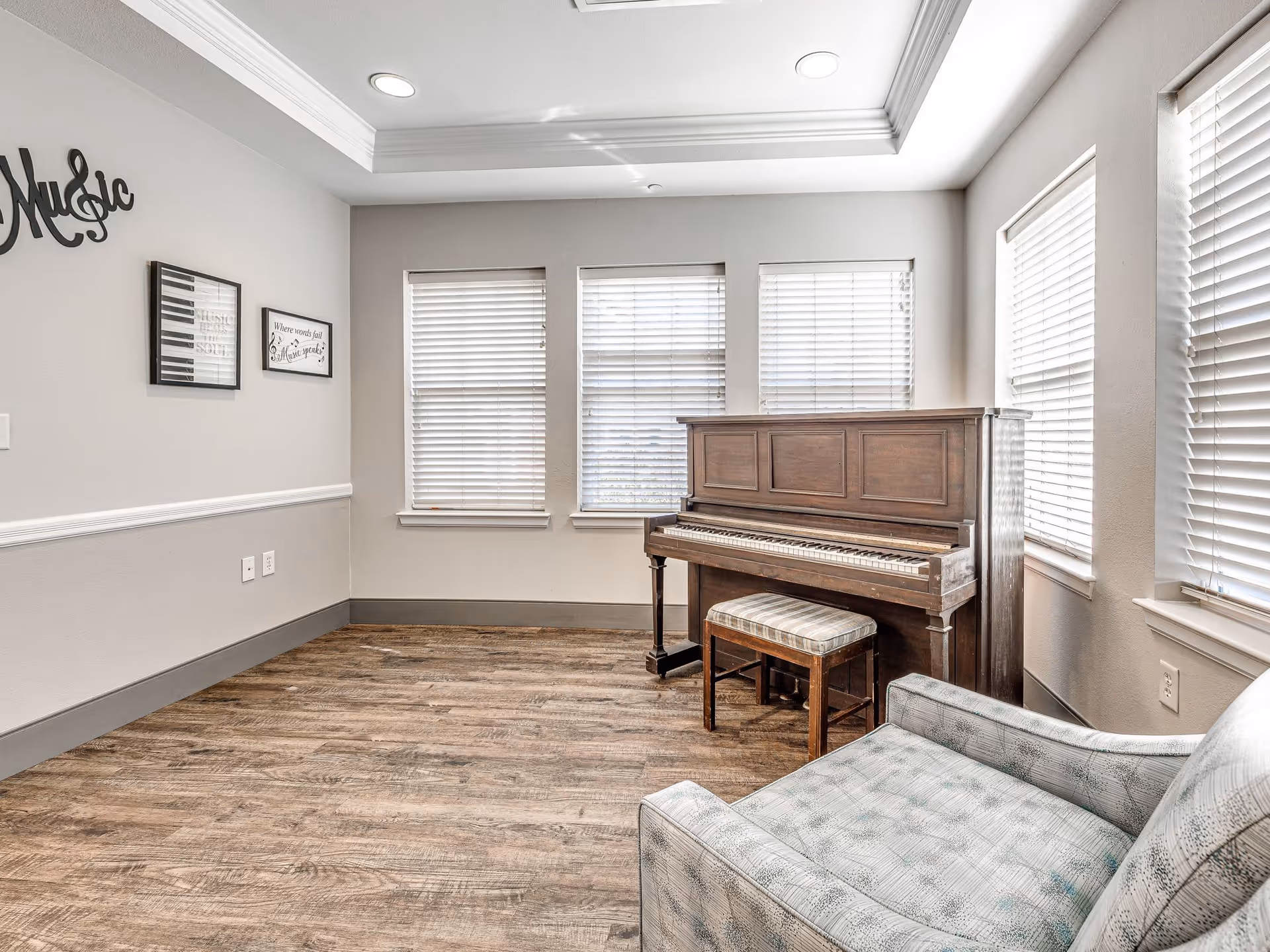 A bright room with wooden flooring featuring an upright piano with a cushioned bench in front of three windows with blinds. A patterned armchair is partially visible in the foreground on the right. The walls are light gray with white trim, and there are framed musical-themed decorations on the left wall.