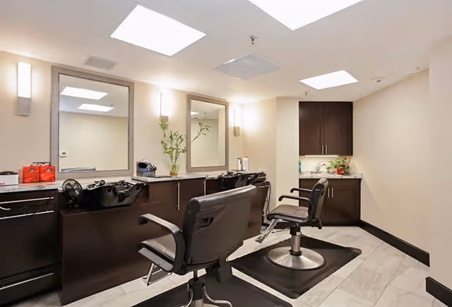 Interior view of a salon area with two black salon chairs in front of two wash basins and large mirrors mounted on the wall. The room has light-colored walls, recessed ceiling lights, and dark wood cabinetry with a small countertop and sink in the corner.