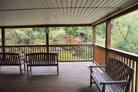 Covered wooden porch with benches and a railing overlooking a wooded area.