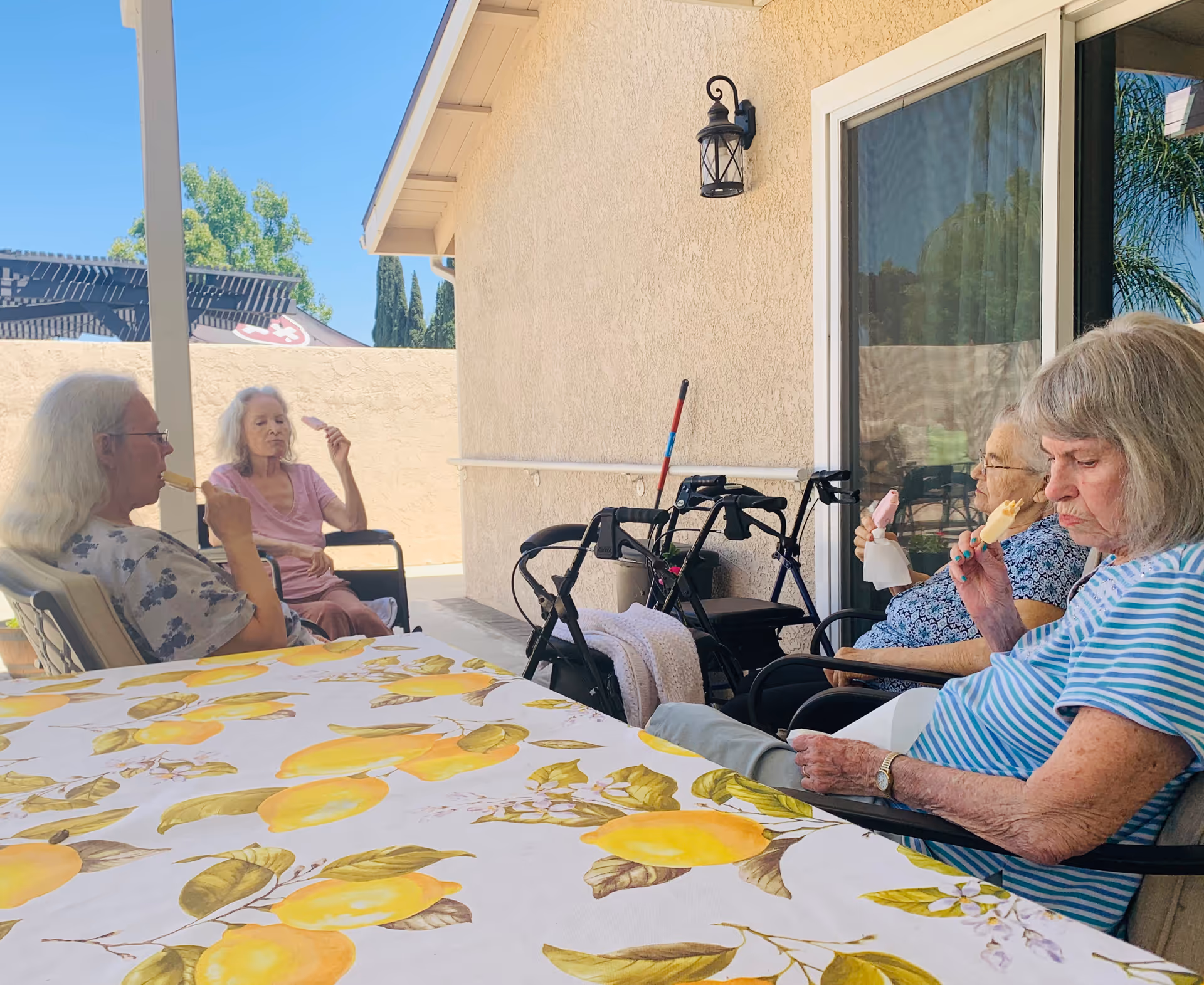 Four elderly women sitting outside on a patio, each enjoying an ice cream bar. They are seated around a table with a lemon-patterned tablecloth. There are walkers and a broom against the wall behind them, and a sliding glass door is visible.