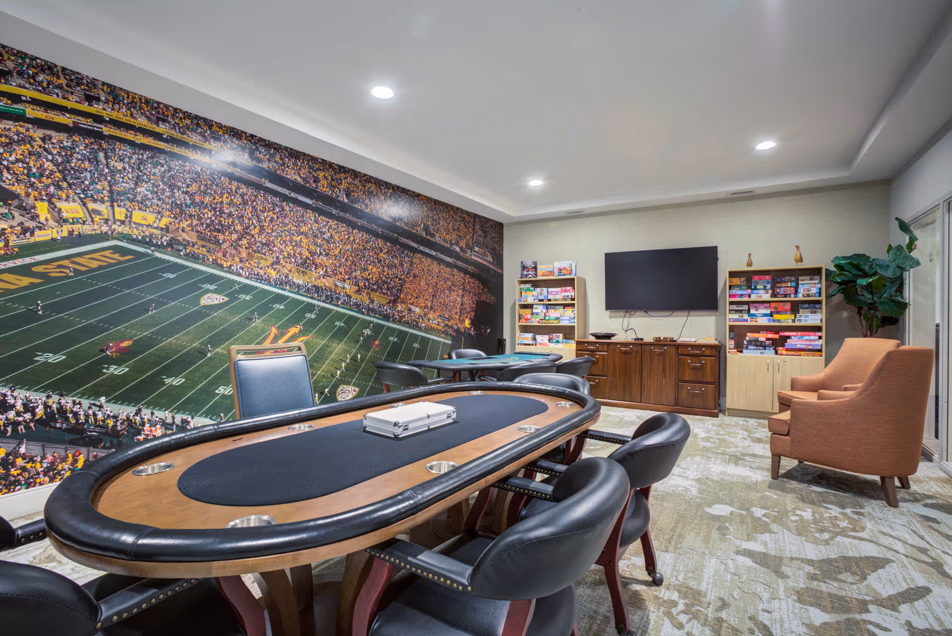 A game room with a poker table surrounded by black leather chairs, a large wall mural of a football stadium, shelves filled with board games, a flat-screen TV mounted on the wall, and an orange armchair next to a large green plant.