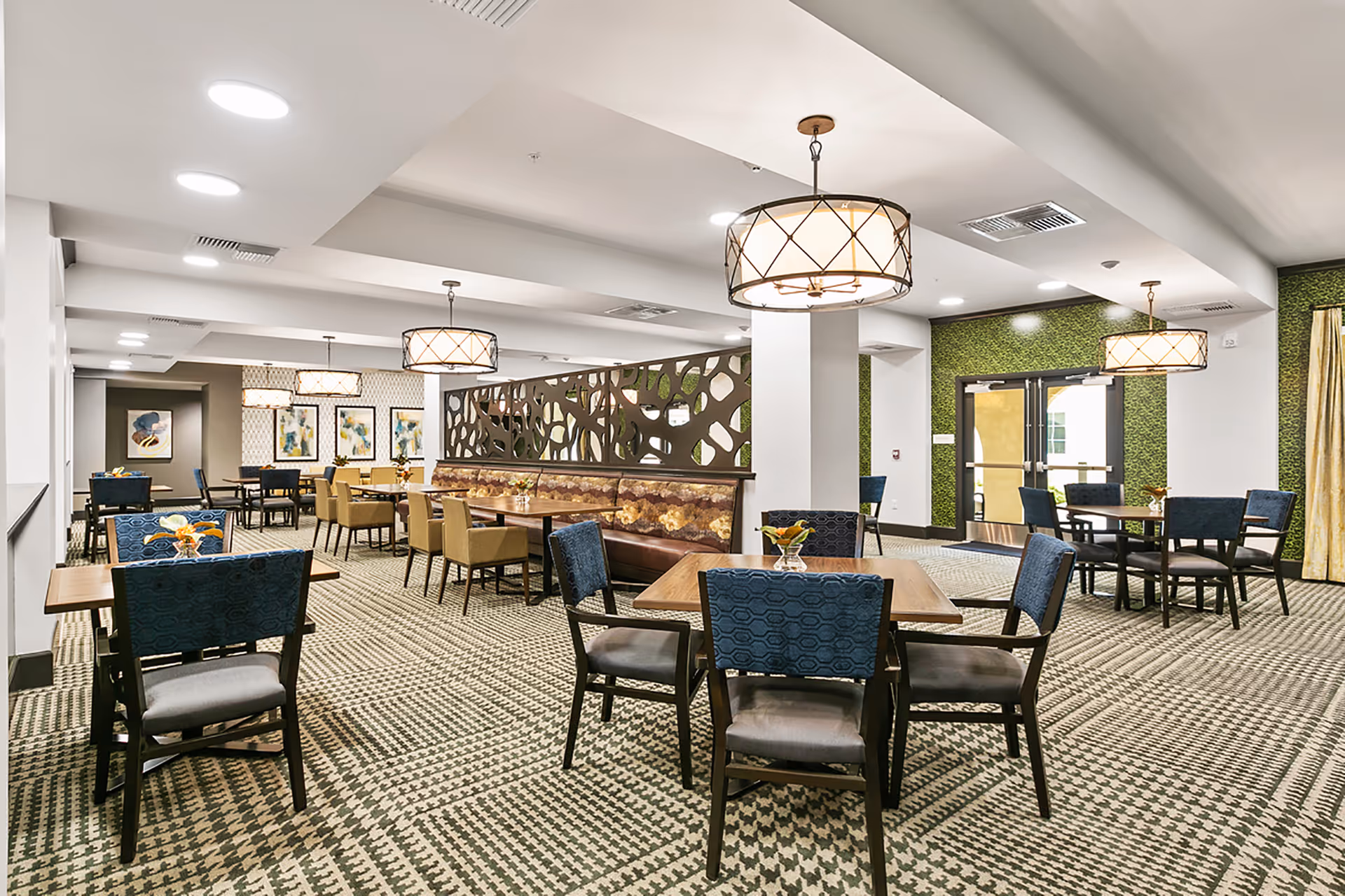 A spacious dining room with multiple tables and chairs arranged neatly. The room features patterned carpet flooring, modern pendant lights hanging from the ceiling, and decorative wall art. There is a partition with an abstract design separating part of the room, and large windows with curtains allowing natural light to enter.