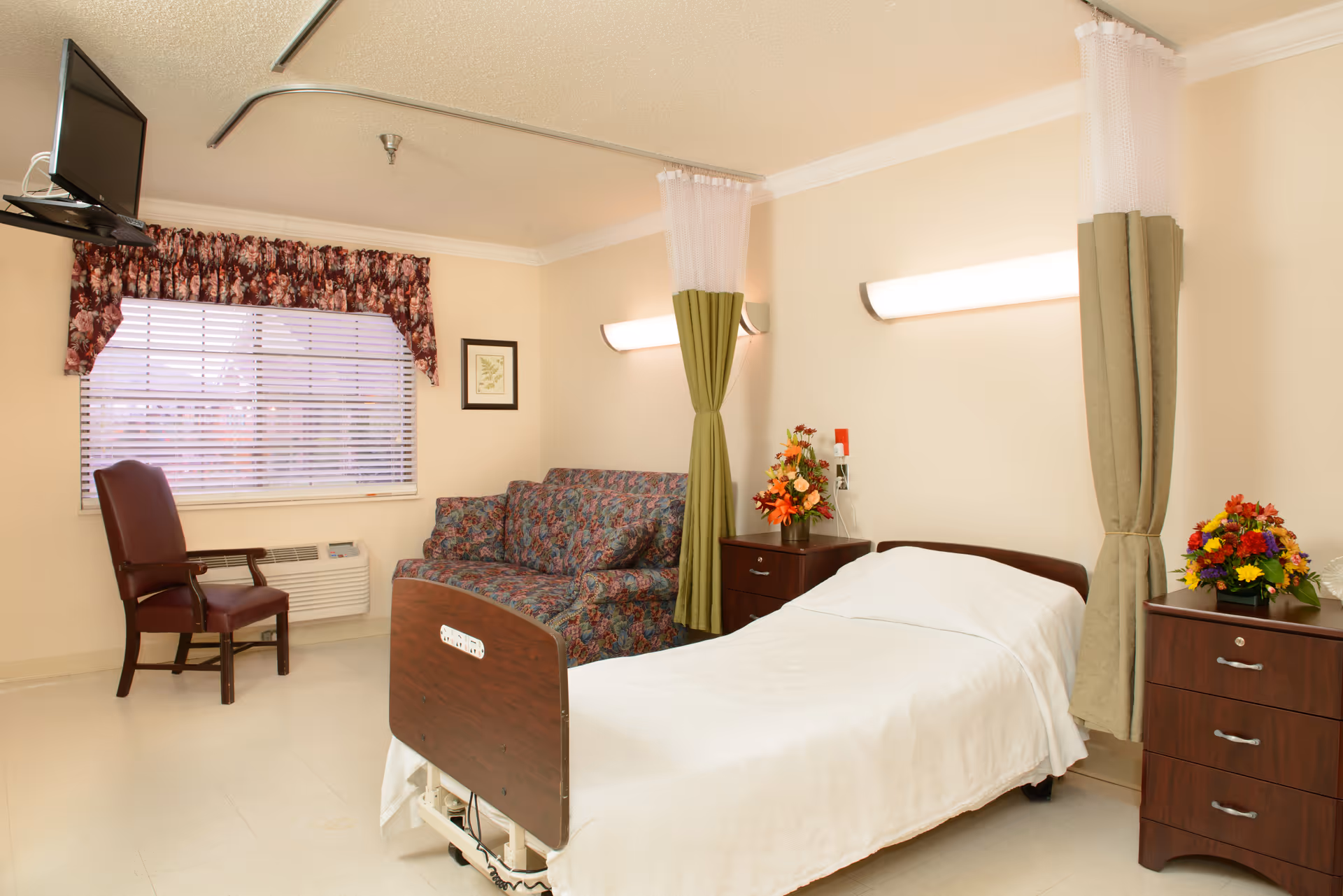 A clean and well-lit patient room in a senior living facility featuring a single hospital bed with white linens, a floral-patterned couch, a wooden chair, two bedside tables with colorful flower arrangements, a window with blinds and floral valance, and a wall-mounted TV in the corner.