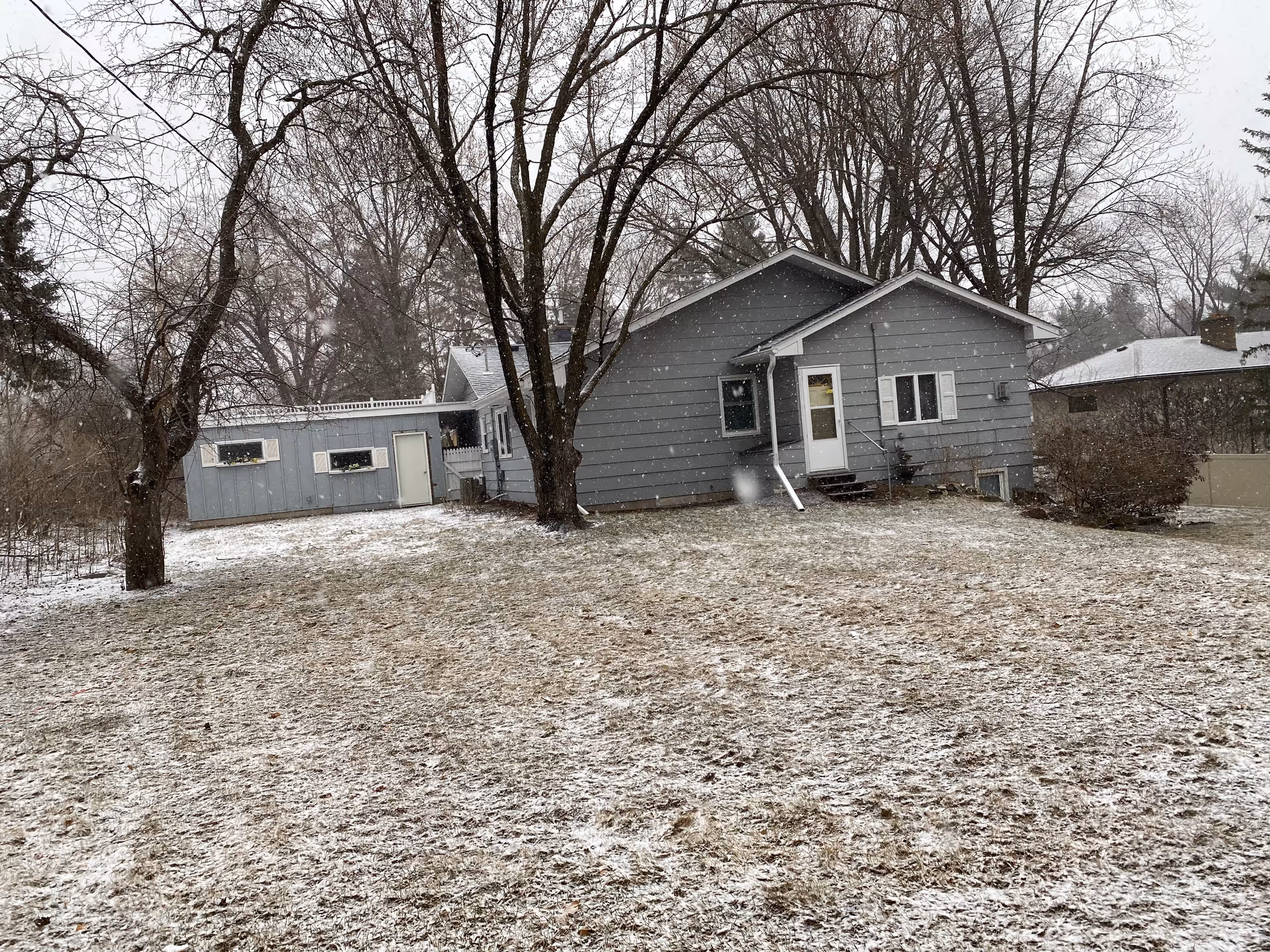 Gray single-story house with an attached shed and bare trees in a lightly snow-covered backyard.