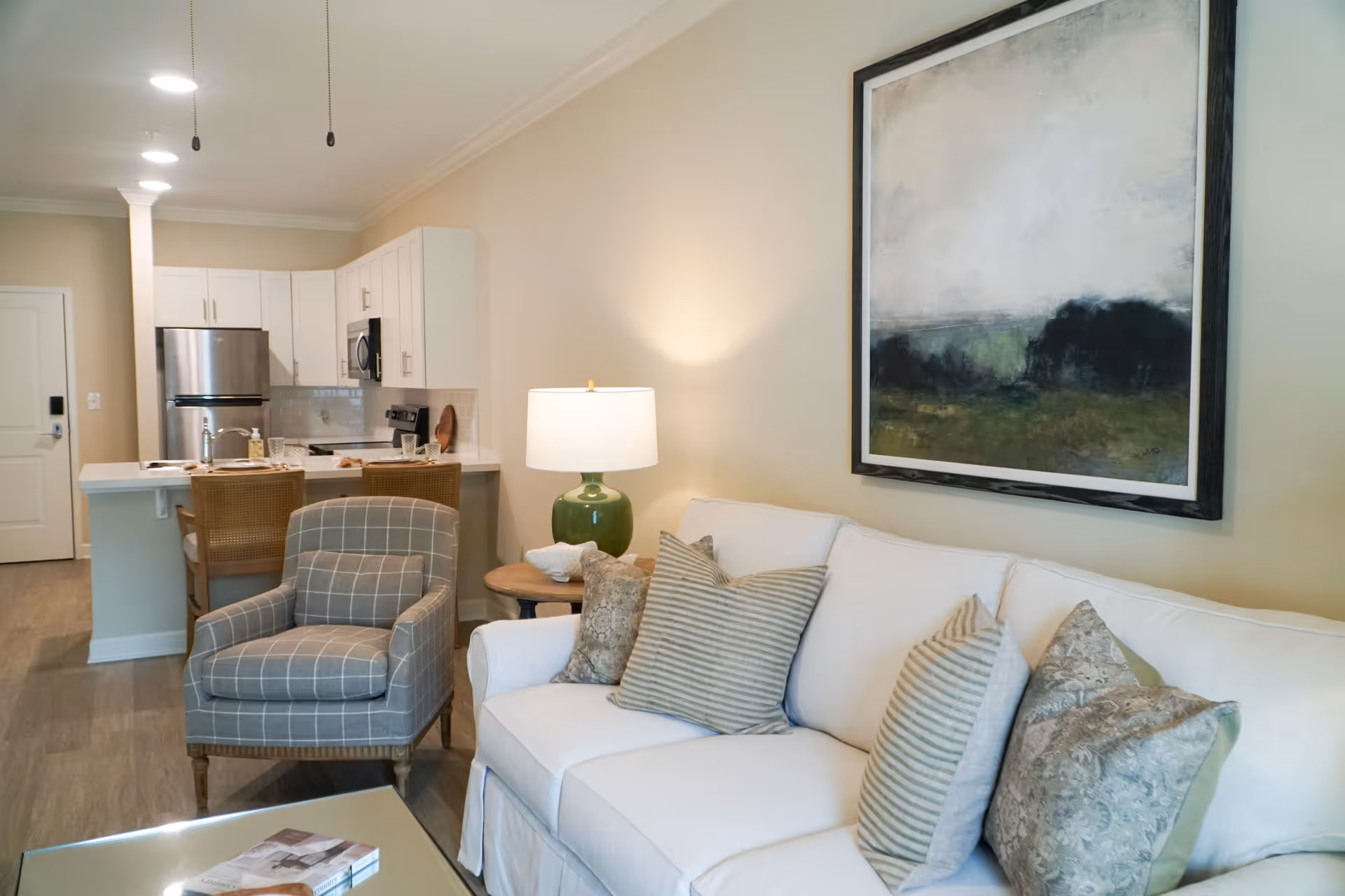 A cozy living room area with a white sofa adorned with multiple patterned pillows, a green table lamp on a wooden side table, and a large framed abstract painting on the wall. In the background, there is a kitchen with white cabinets, a stainless steel refrigerator, microwave, and stove, along with a breakfast bar with two wicker chairs.
