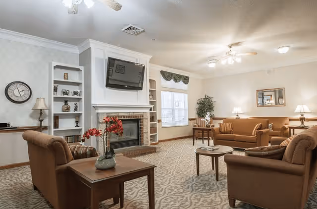 A cozy living room with beige walls and carpet, featuring a brick fireplace with a mounted flat-screen TV above it. The room has multiple brown upholstered chairs and sofas arranged around wooden coffee and side tables. There are decorative items on built-in shelves next to the fireplace, a wall clock, table lamps, a large window with a green valance, and a framed mirror on the wall.