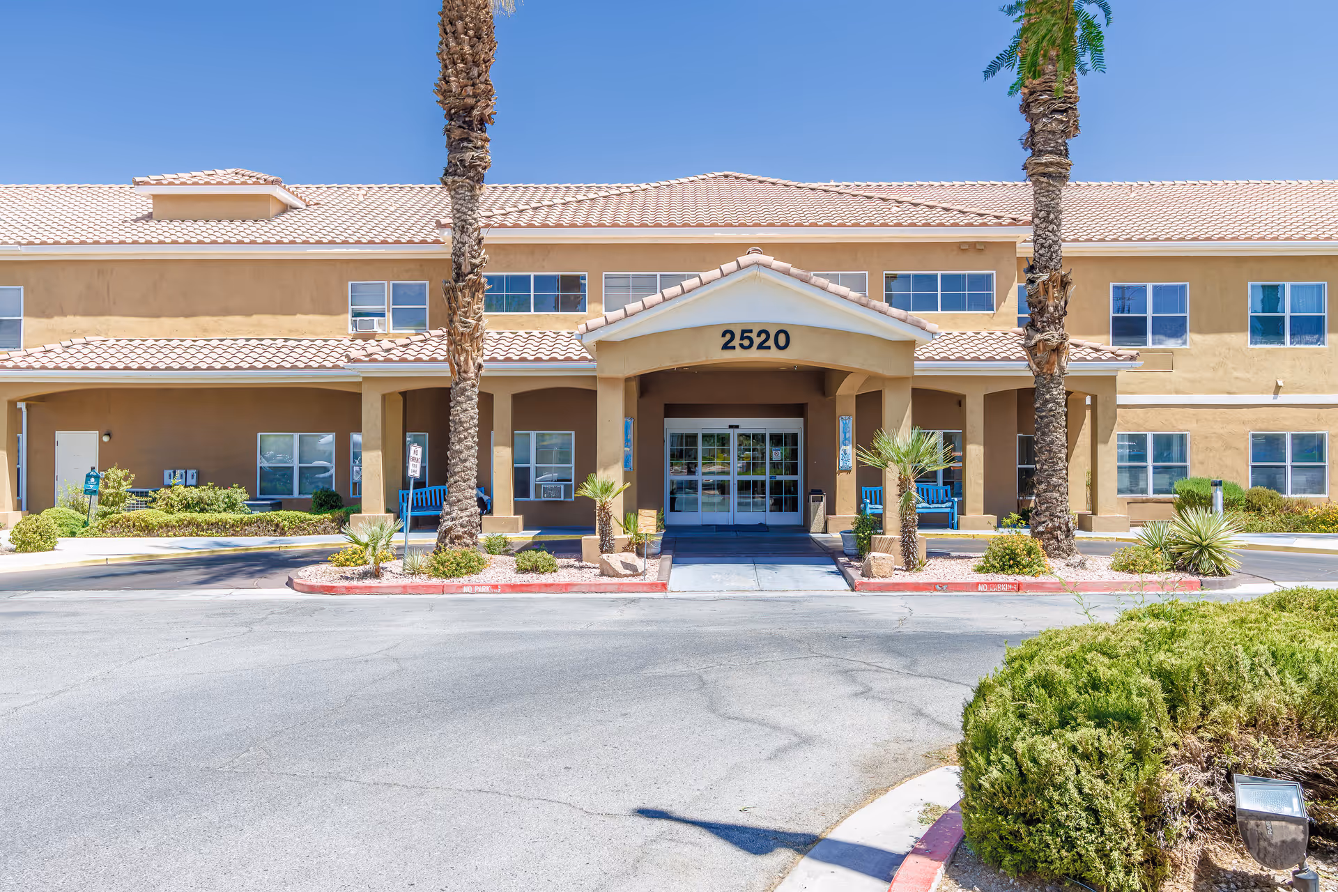 Front exterior view of a two-story senior living facility building with beige walls and a tiled roof. The entrance has a covered porch with the number 2520 above it, flanked by palm trees and blue benches. There are shrubs and desert landscaping around the driveway and entrance area under a clear blue sky.