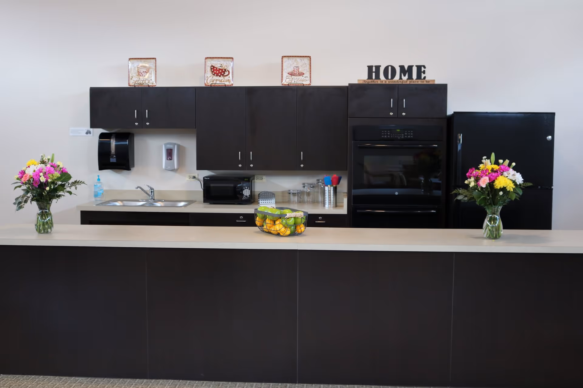 Modern kitchenette with dark cabinets, a long counter holding two vases of flowers and a fruit bowl, sink, microwave, oven and refrigerator.