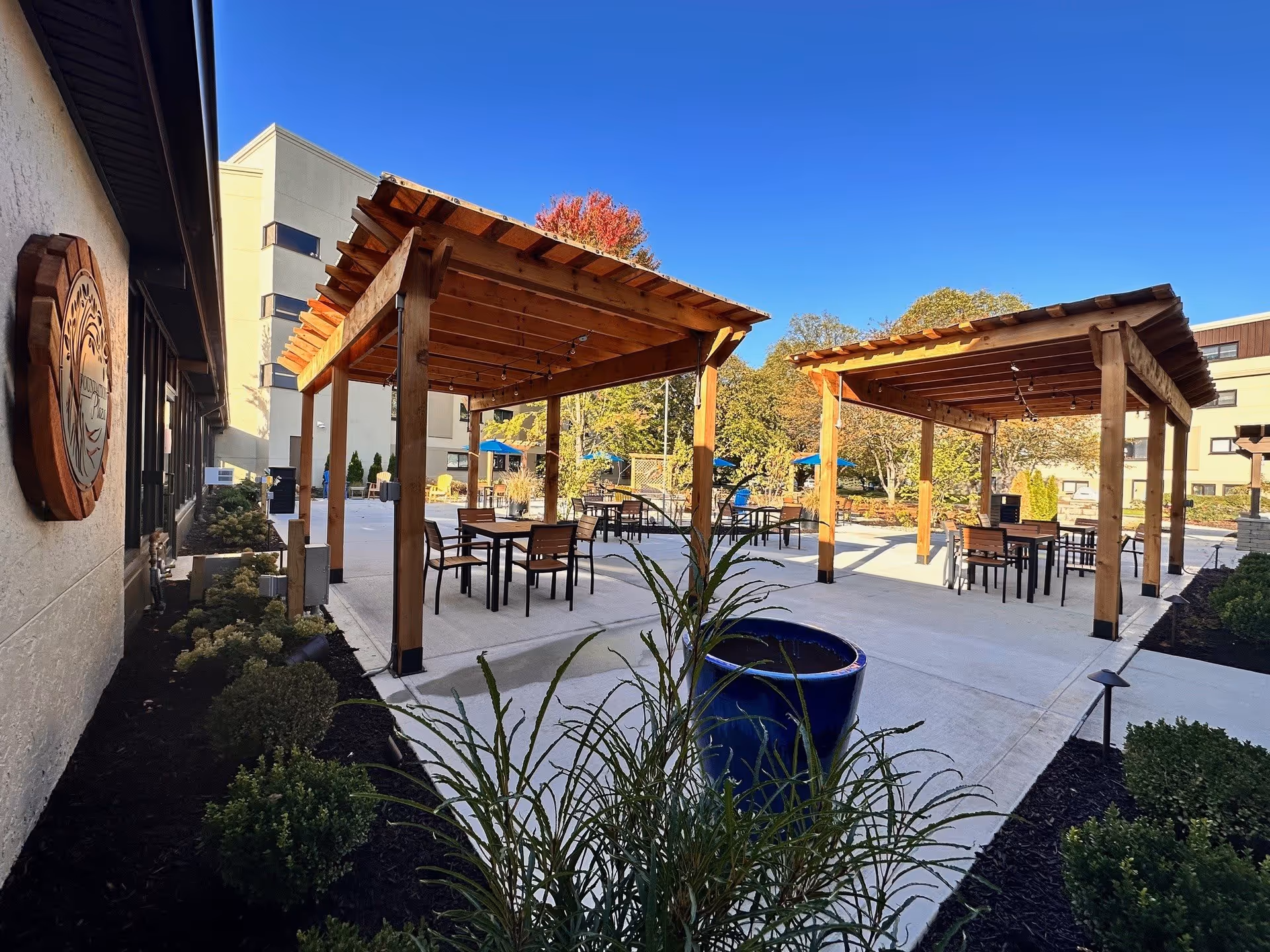 Outdoor patio area at Westminster Village featuring two wooden pergolas with string lights, several tables and chairs underneath, surrounded by landscaped bushes and plants, with a clear blue sky and building structures in the background.