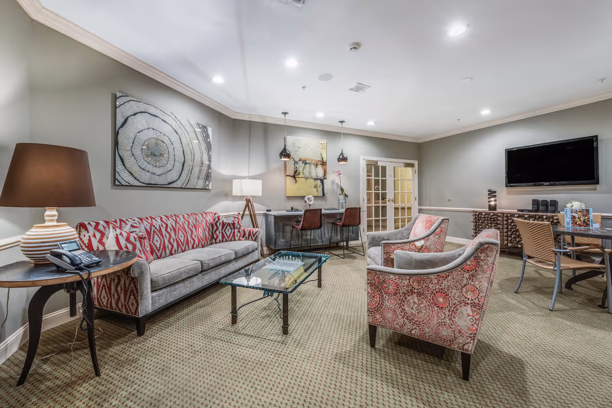 A cozy living room area with a patterned red and gray sofa, two matching armchairs, a glass coffee table with a chess set, a side table with a lamp and telephone, a desk with two chairs and pendant lights, a wall-mounted TV, and decorative artwork on the walls.