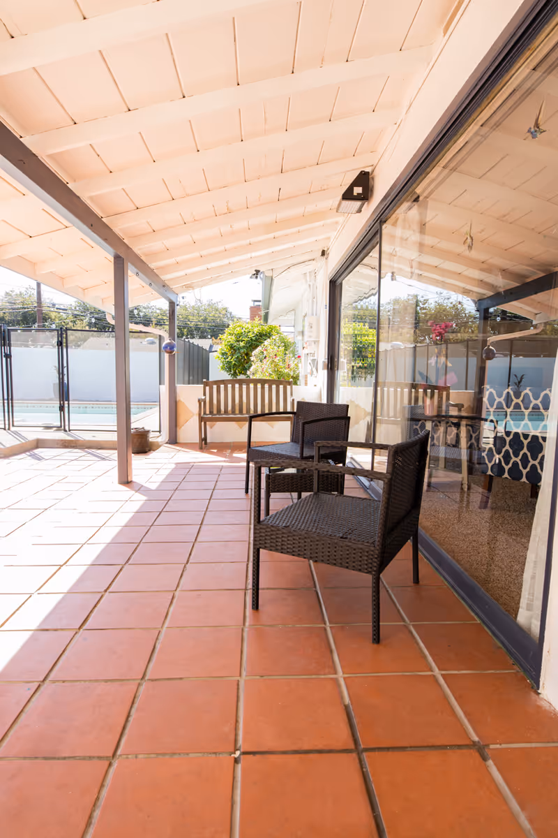 Covered outdoor patio area with terracotta tile flooring, two black wicker chairs, a wooden bench, and a glass sliding door reflecting the patio furniture and interior. There is a pool fenced in the background and some greenery near the bench.