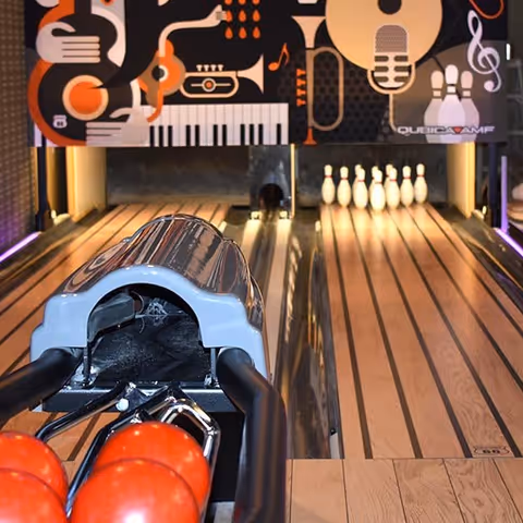 View down a bowling lane with red bowling balls in the foreground and pins set up at the end of the lane. Above the pins is a colorful graphic featuring musical instruments and a microphone.