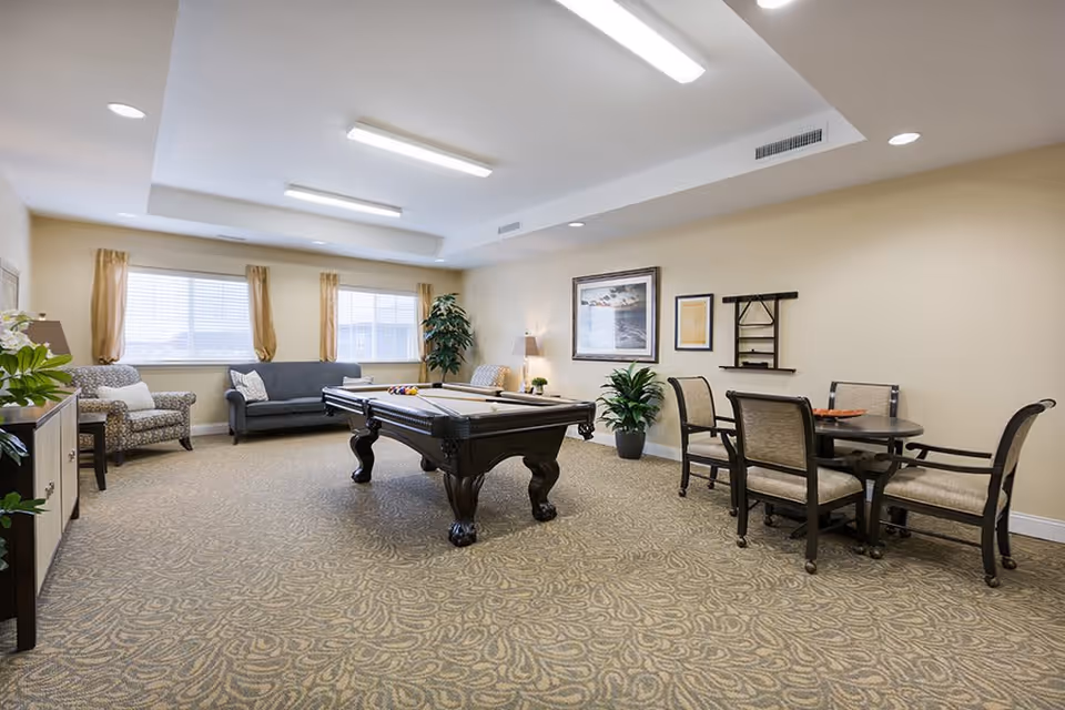 A spacious common room with a pool table in the center, surrounded by seating including a gray sofa, two patterned armchairs, and a round table with four chairs. The room has beige walls, patterned carpet, large windows with light brown curtains, and framed artwork on the walls. There are also several potted plants and ceiling lights providing illumination.