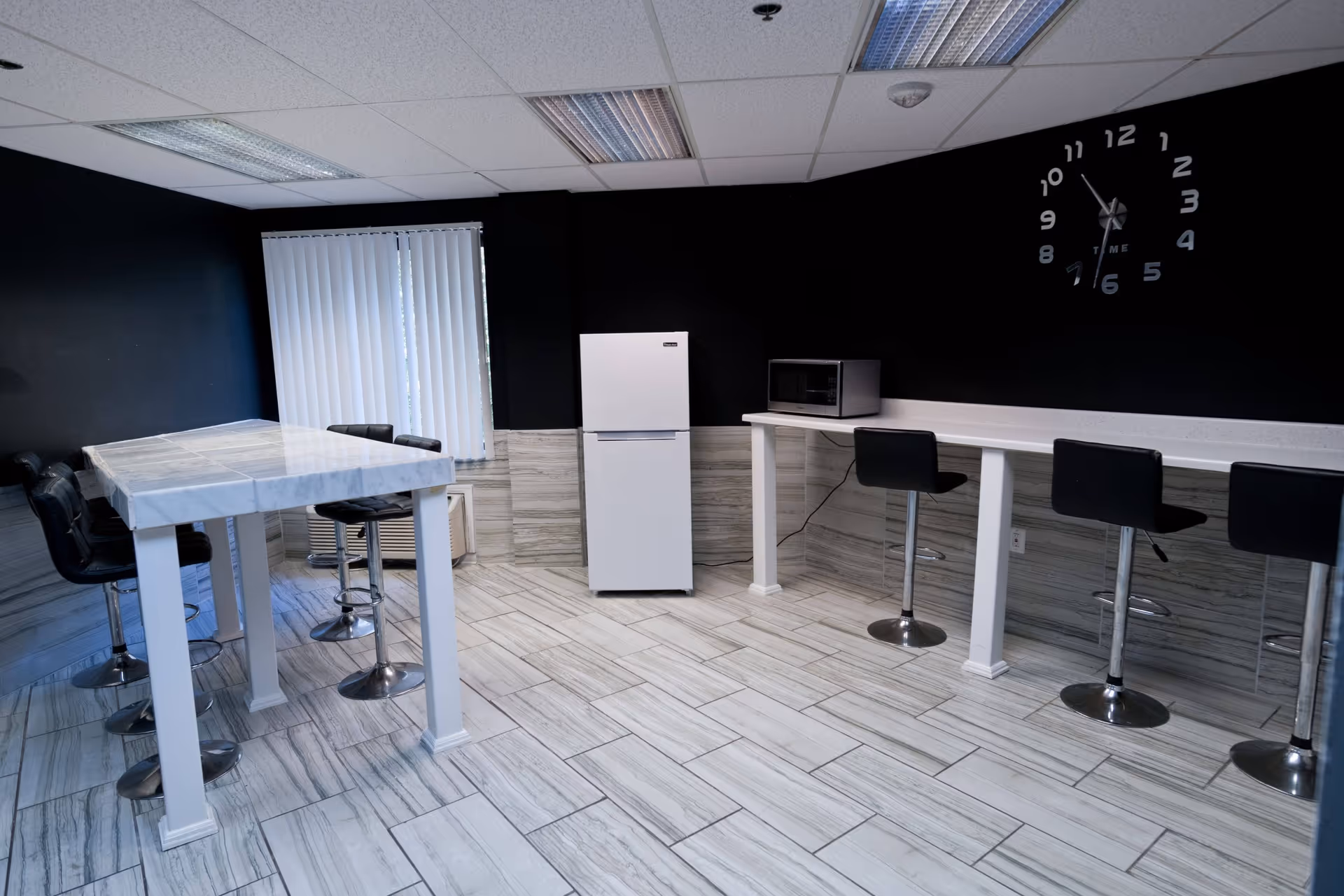 A modern kitchen area with a white refrigerator, a microwave on a long white counter, and black bar stools. There is a high table with several black chairs on the left side near a window with vertical blinds. The floor is tiled with light-colored tiles, and the walls are painted black with a large wall clock displaying the time.