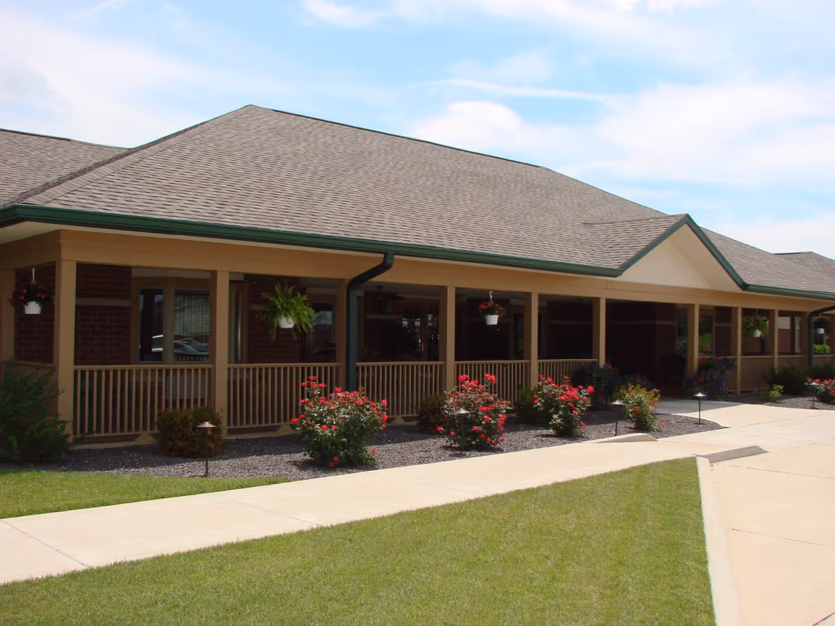 Exterior view of a single-story building with a sloped roof, a covered porch with railings, hanging plants, and flower beds with red flowers along the walkway.