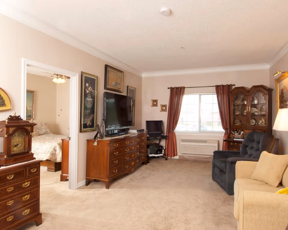 A cozy living room with beige walls and carpet, featuring a dark blue armchair, a beige sofa, a wooden dresser with a flat-screen TV on top, a wooden display cabinet with glass doors, and a window with red curtains. An adjacent bedroom with a bed and ceiling fan is visible through an open doorway.