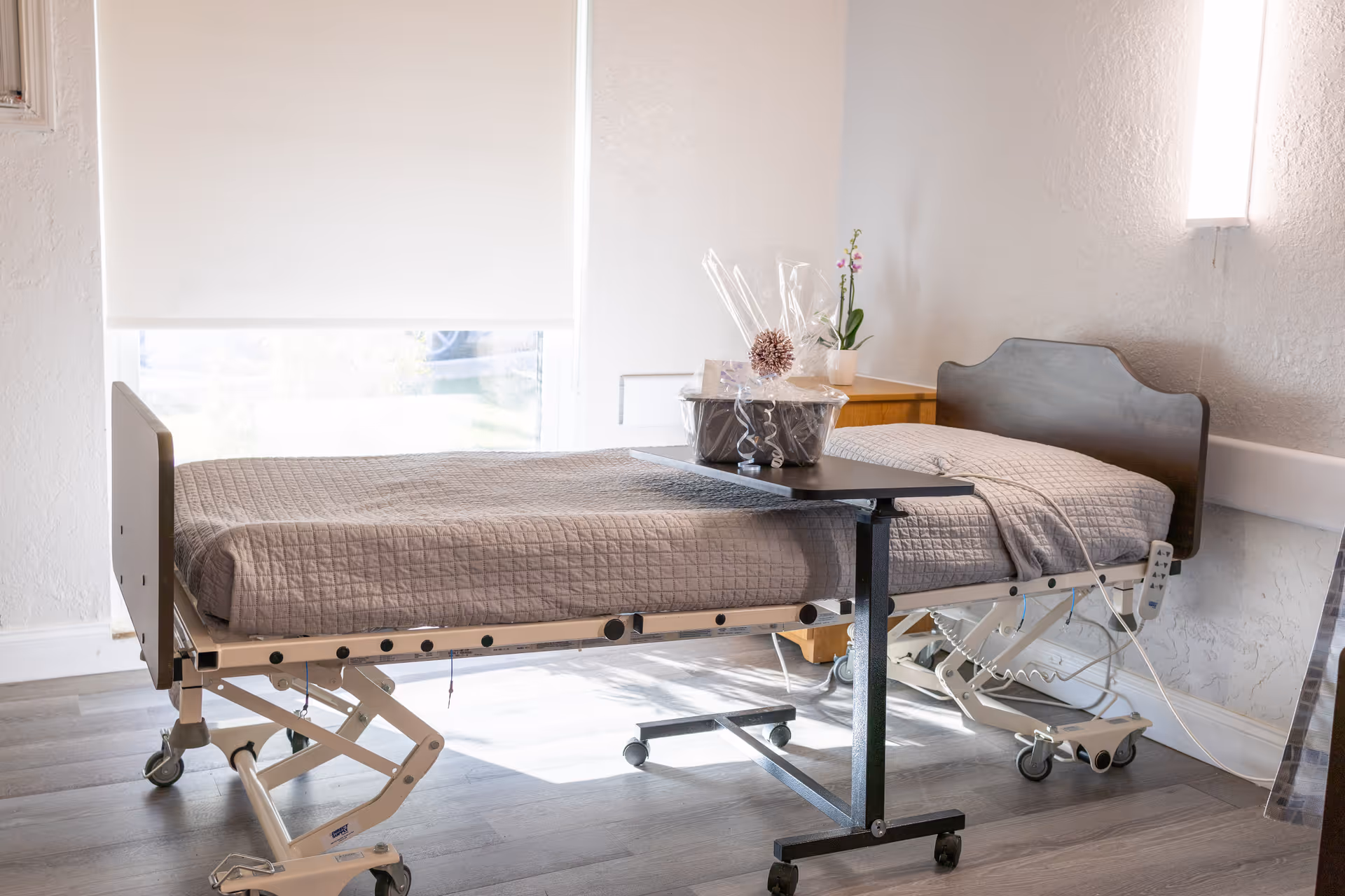 A hospital-style bed with a gray quilted blanket in a bright room with a large window covered by a white roller shade. Next to the bed is a small wooden nightstand with a potted plant and a gift basket on an overbed table. The floor is wood, and a modern wall light is mounted on the wall above the bed.