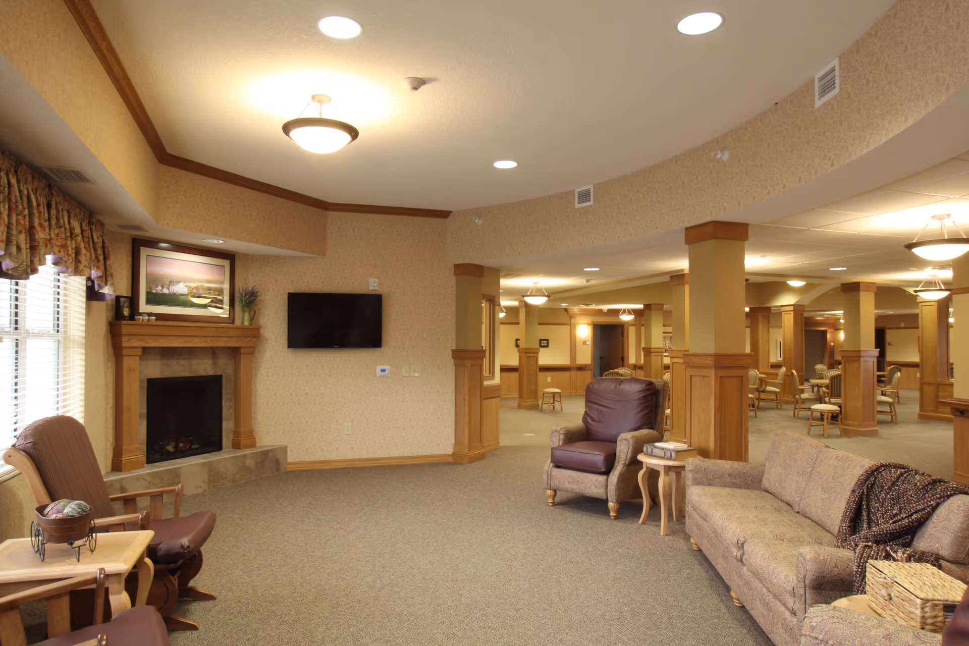A spacious and warmly lit common area in a senior living facility featuring a fireplace with a framed picture above it, a wall-mounted TV, comfortable seating including armchairs and a sofa with a throw blanket, wooden pillars, and several tables and chairs in the background.