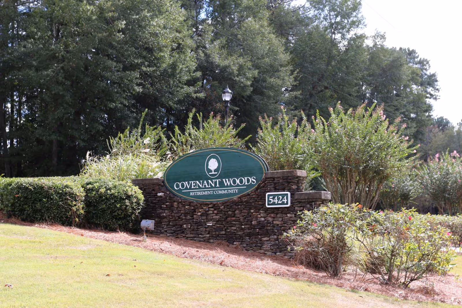Stone sign at the entrance of Covenant Woods retirement community surrounded by greenery and bushes with trees in the background.