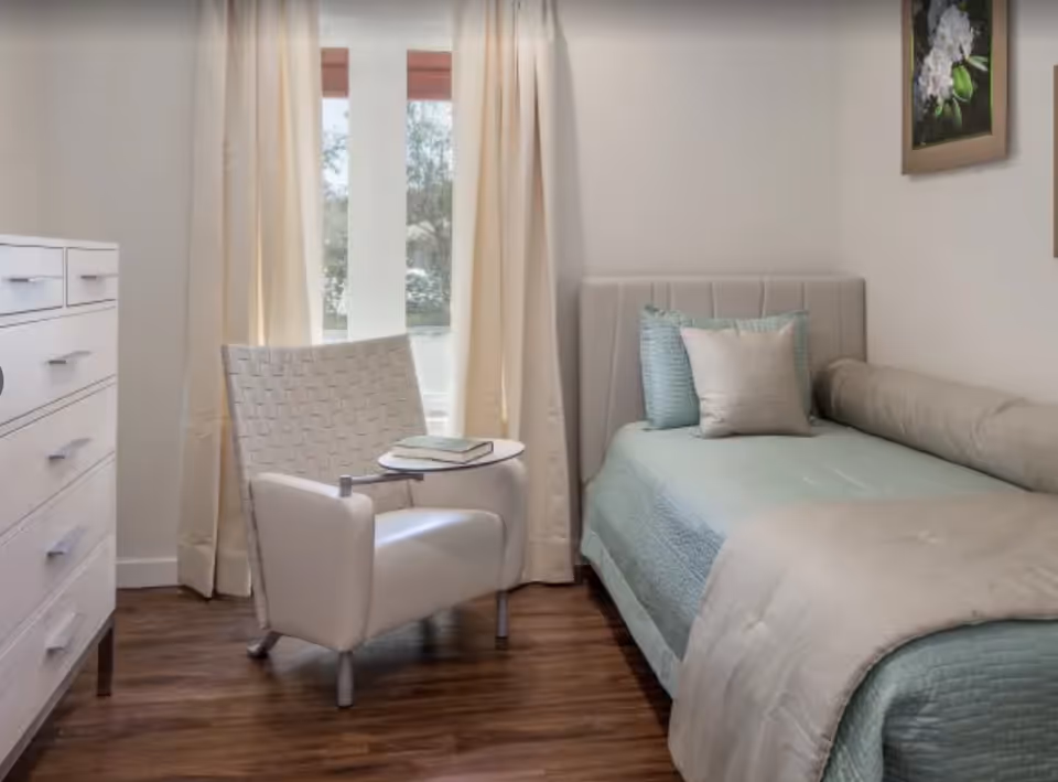 A small bedroom with a single bed covered in light blue and beige bedding, two decorative pillows, and a bolster pillow. Next to the bed is a white armchair with a woven backrest and a small attached round table holding a closed book. A white dresser with multiple drawers is on the left side, and beige curtains cover a window behind the chair. The floor is wooden, and a framed floral picture hangs on the wall above the bed.