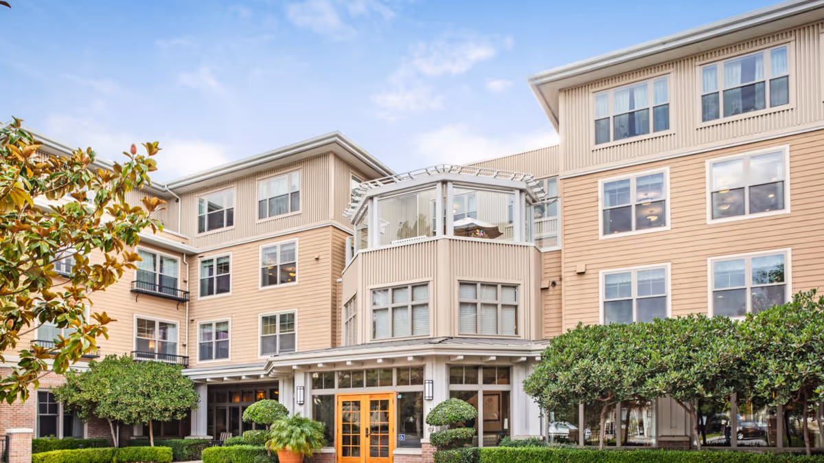 Front exterior of a multi-story beige senior living building with large windows, a central entrance and manicured shrubs.