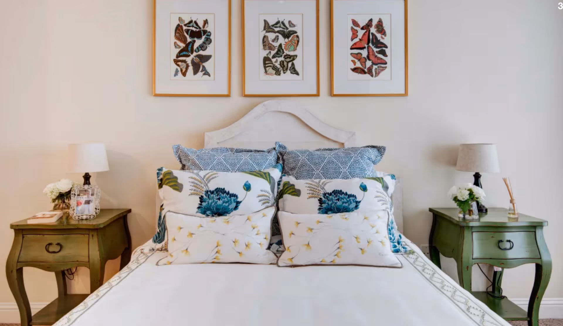 A neatly made bed with a white headboard and multiple decorative pillows featuring blue and floral patterns. On each side of the bed are green wooden nightstands with lamps, flower arrangements, and a framed photo on the left nightstand. Above the bed are three framed butterfly artworks.