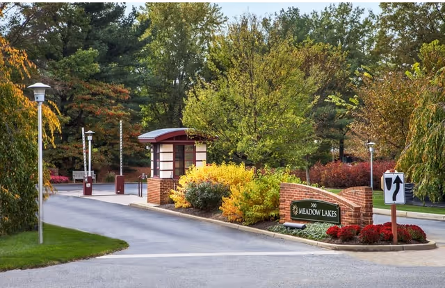 Entrance to Meadow Lakes facility with a small guardhouse, brick walls, colorful bushes, trees, and a road with a traffic sign indicating a left or right turn.