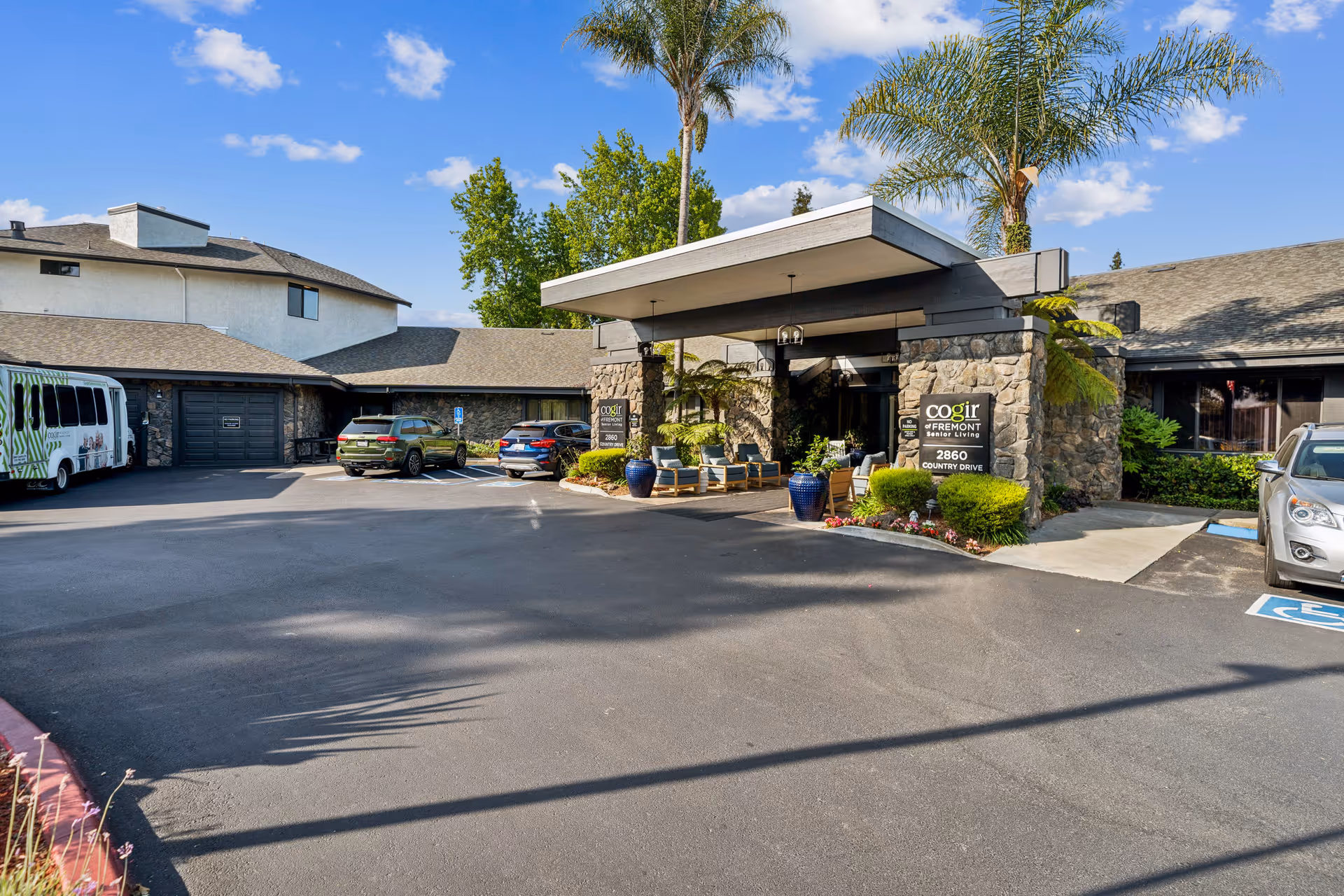 Front entrance and covered drop-off of the Cogir of Fremont senior living facility with parked cars, potted plants, and palm trees.