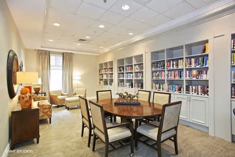 A well-lit interior room with a round wooden table surrounded by six upholstered chairs. Along one wall, there are built-in bookshelves filled with books. In the background, there are two armchairs and a floor lamp near a window with curtains. The room has a beige carpet and a ceiling with recessed lighting.