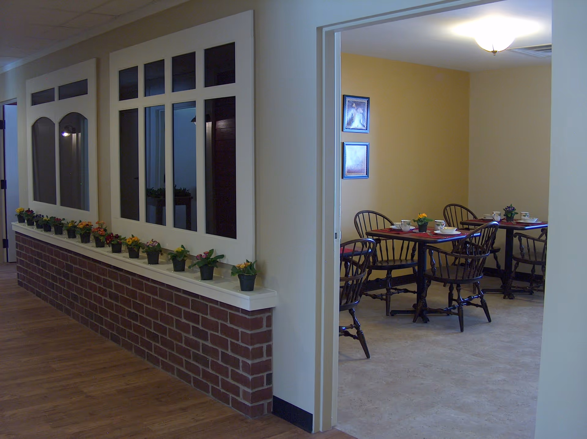 Interior view of a senior living facility hallway with a brick and white half-wall adorned with small potted flowers. Through an open doorway, a dining area is visible with tables set with cups and plates, surrounded by wooden chairs. The walls are painted yellow and decorated with framed pictures.