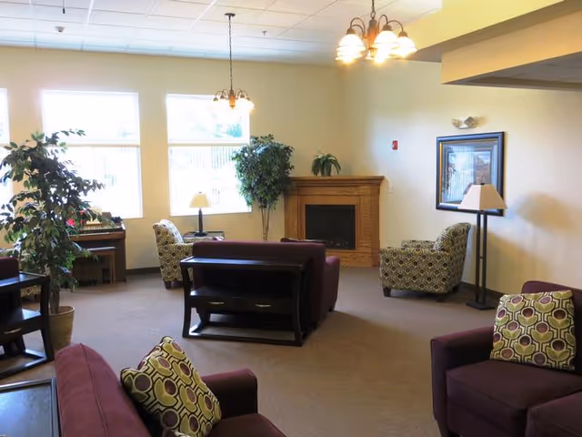 A cozy living room area with multiple armchairs and sofas upholstered in purple fabric, some with patterned cushions. There is a wooden fireplace with a plant on top, a framed picture on the wall, and several lamps providing warm lighting. Large windows let in natural light, and there are a couple of potted plants adding greenery to the space.