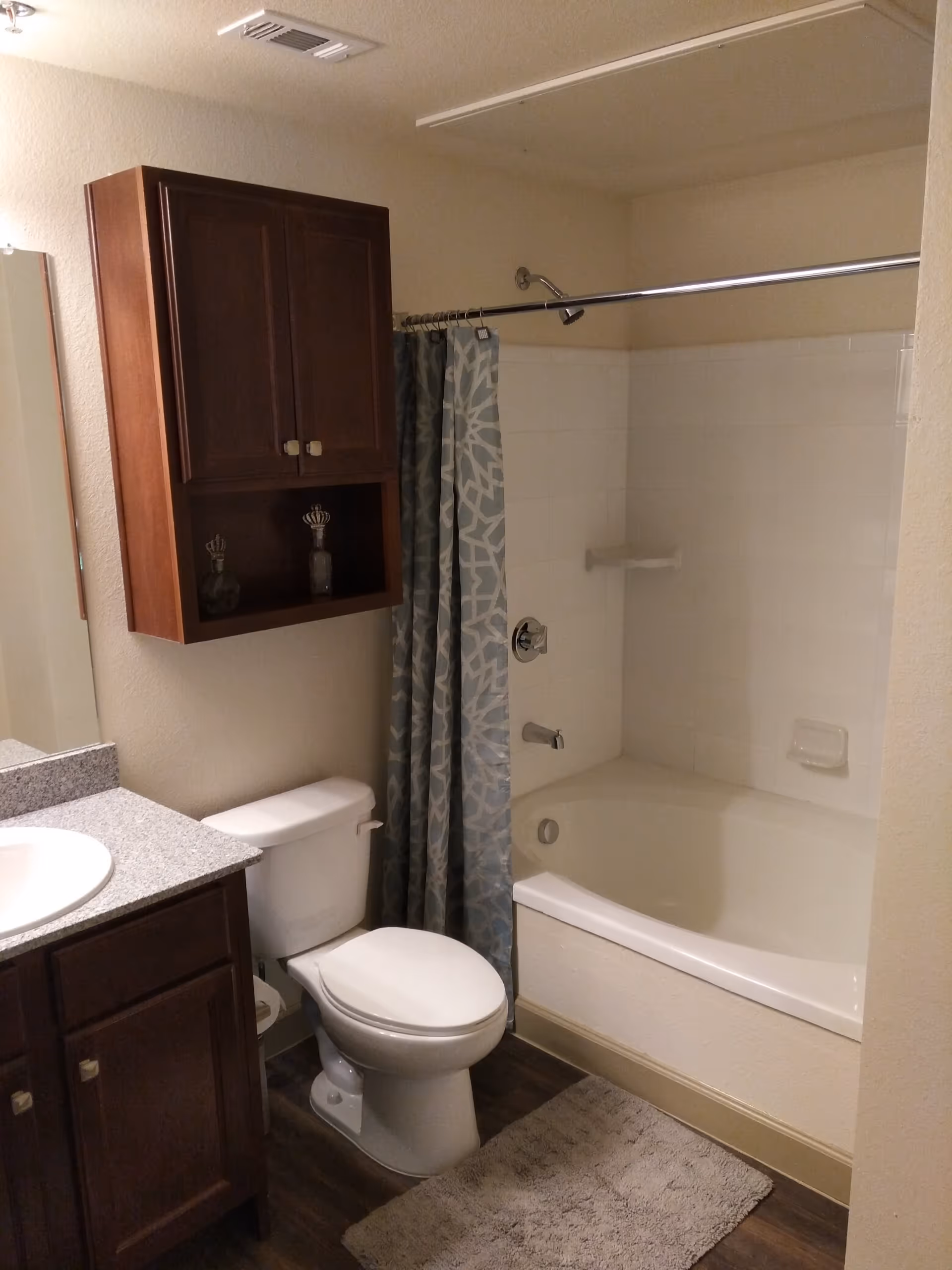 A bathroom featuring a white toilet, a bathtub with a shower and a blue patterned shower curtain, a wooden cabinet mounted on the wall above the toilet, and a countertop with a sink. The floor has a dark wood finish and there is a light-colored bath mat in front of the bathtub.