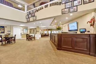 Spacious senior living facility interior with a large reception desk on the right, a 'Wall of Honor' display with framed photos on the upper wall, multiple seating areas with tables and chairs, and a carpeted floor. The area is well-lit with ceiling lights and decorated with flowers and artwork.