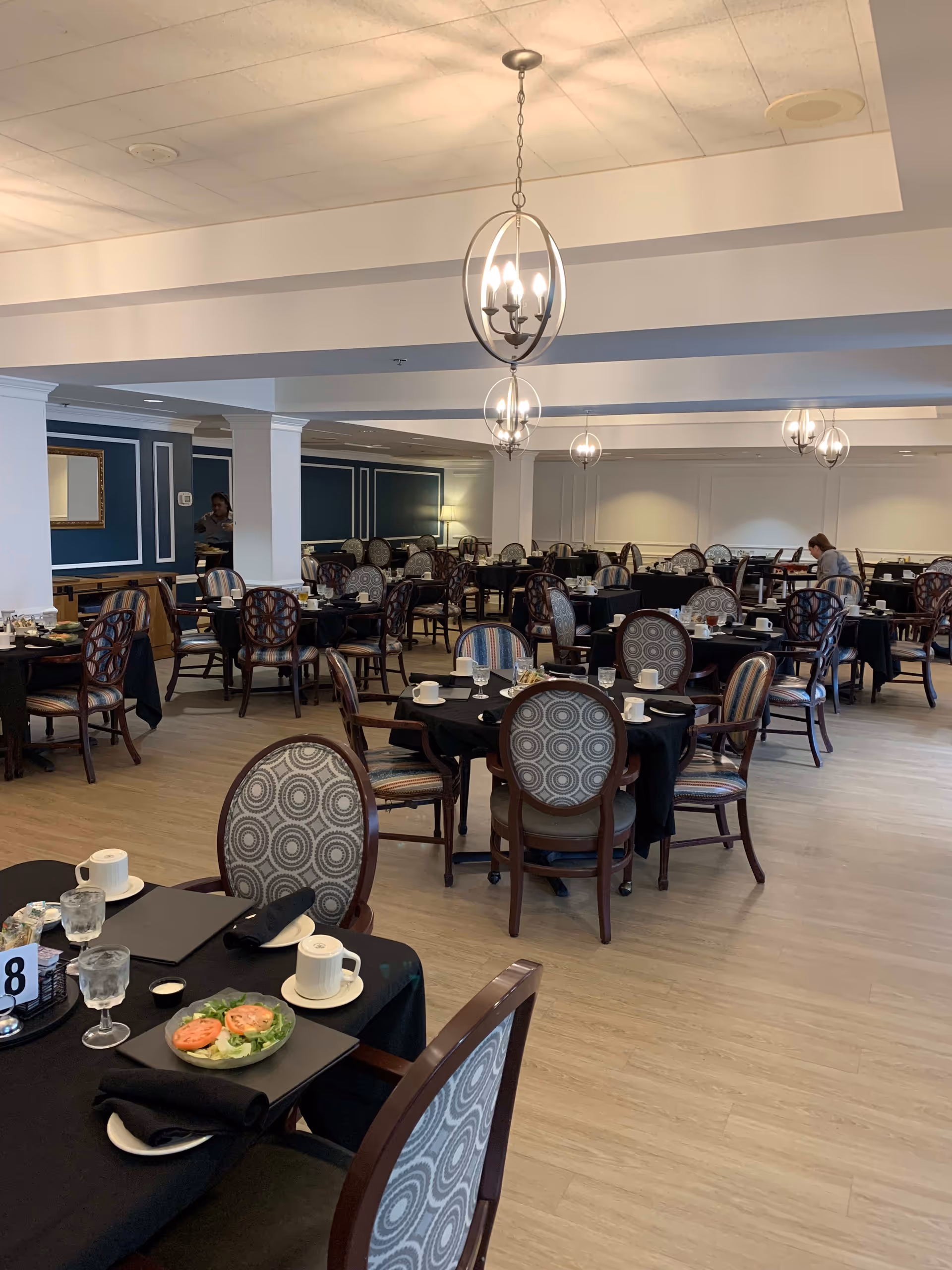 A spacious dining room with multiple round tables covered with black tablecloths, each set with cups, glasses, plates, and napkins. The chairs have patterned upholstery, and modern spherical chandeliers hang from the ceiling. A few people are seated or standing in the background.
