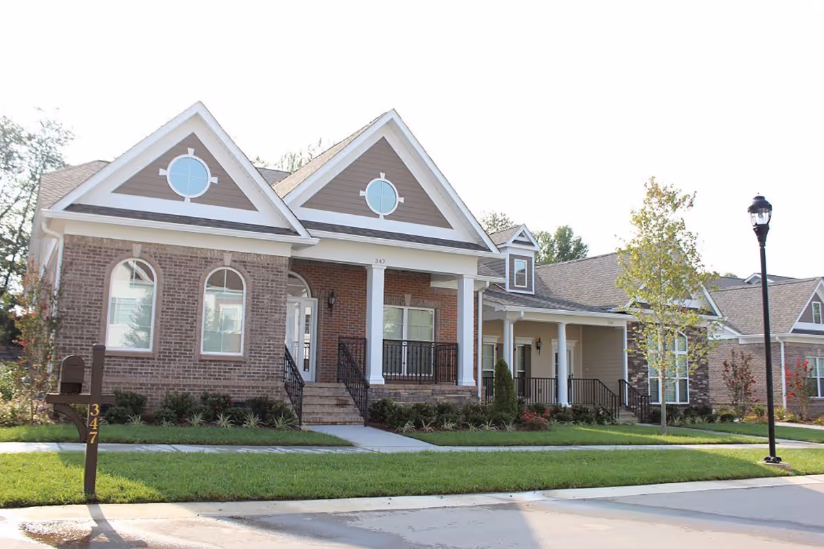 Exterior view of a brick building with a peaked roof and large windows, featuring a front porch and landscaped yard.