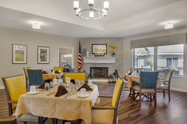 Well-lit dining room with round tables covered in tablecloths set for meals, upholstered chairs, a fireplace, and large windows.