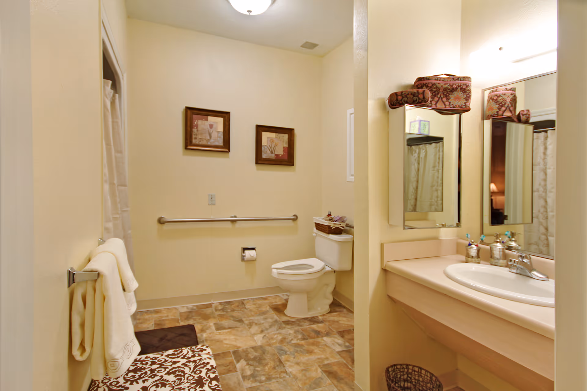 A clean and well-lit bathroom with beige walls and tiled floor. The room features a toilet with a grab bar on the wall beside it, a towel rack with folded towels, a sink with a large mirror above it, and decorative framed pictures on the wall. There is a patterned bench near the towel rack and a basket on the floor under the sink.