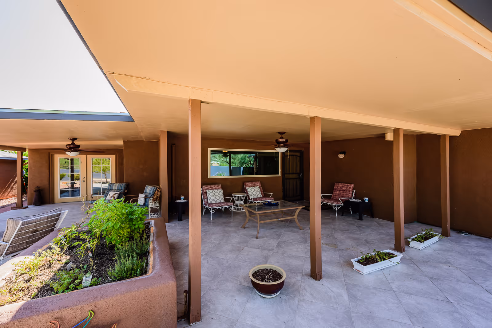 Covered outdoor patio area with several cushioned chairs and a glass coffee table. The patio has a tiled floor, ceiling fans, and is supported by multiple brown pillars. There are planter boxes with greenery and a small garden bed adjacent to the patio.