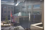 Exterior view of a multi-story building with a covered entrance and three flagpoles displaying flags in front. The building has multiple windows and a landscaped area with small trees and shrubs near the entrance.