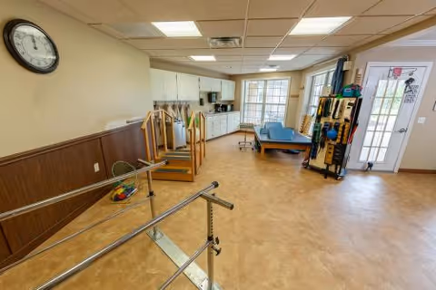 Rehabilitation room with parallel bars, treatment table, exercise equipment, and a kitchenette area in a senior facility.