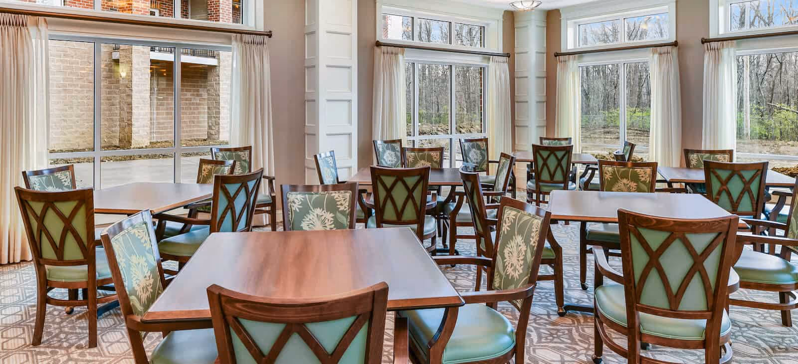 Spacious dining room with wooden tables, green-upholstered patterned chairs, and large windows overlooking wooded grounds.