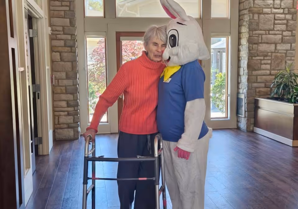 An elderly woman using a walker is hugging a person dressed in a large white bunny costume with a blue shirt and yellow scarf inside a building with stone walls and large windows.