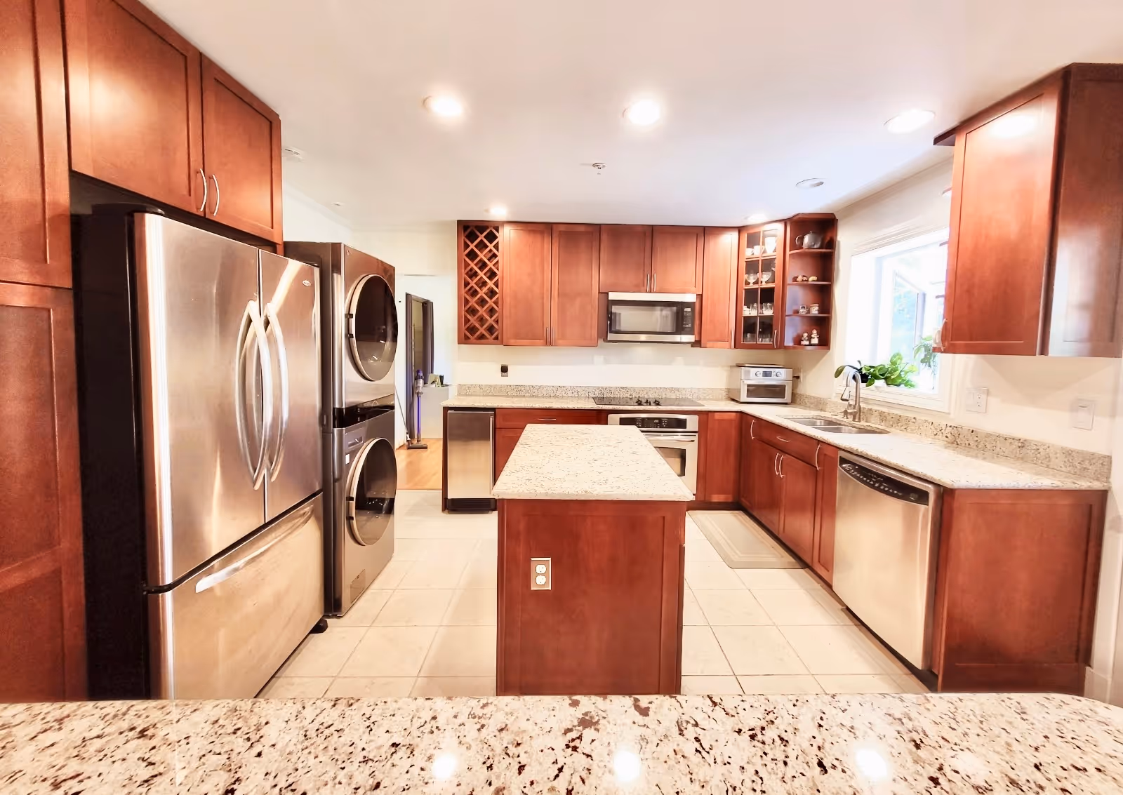 A modern kitchen with cherry wood cabinets, granite countertops, and stainless steel appliances including a refrigerator, dishwasher, microwave, oven, and a stacked washer and dryer. There is a kitchen island in the center and a window above the sink letting in natural light.
